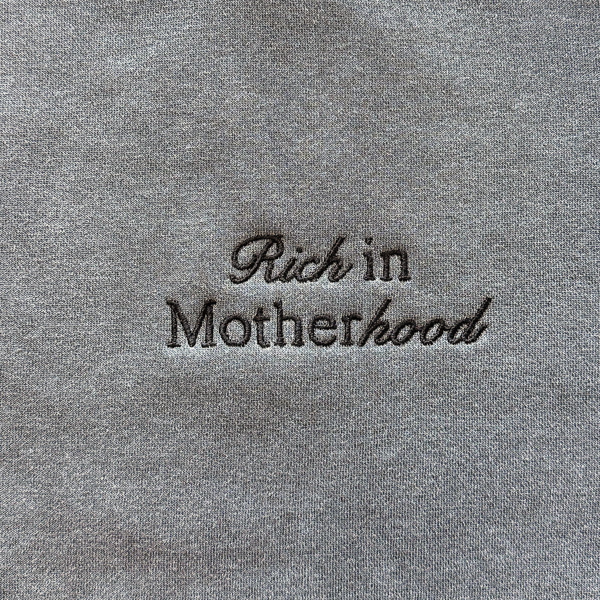 Close-up of the black Rich in Motherhood embroidery on the pepper grey crewneck