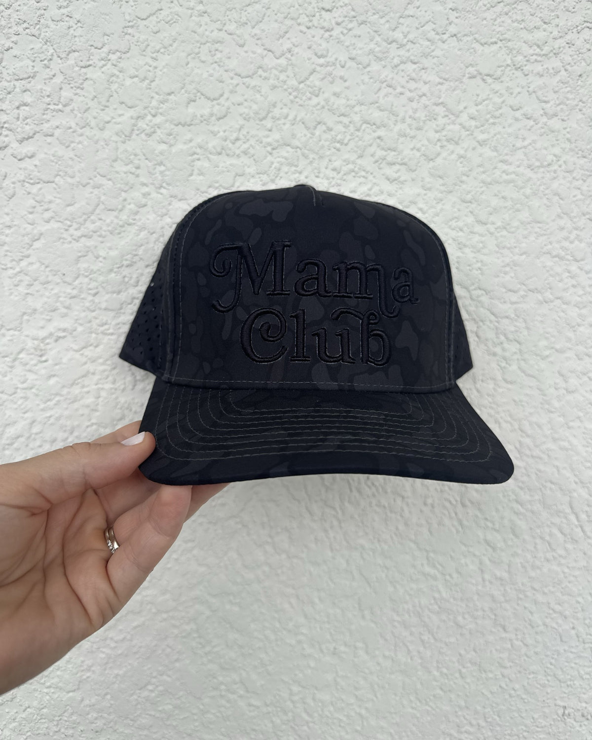 Mama Club black camo trucker, black puff embroidery, held against a white wall